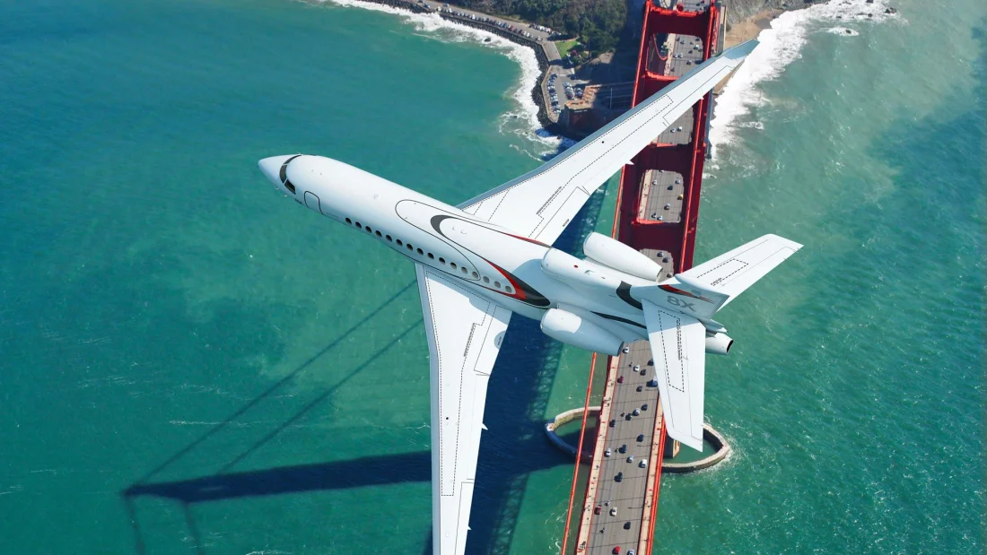 A white Falcon 8X business jet banking low over the Golden Gate Bridge, its shadow on the turquoise sea below.